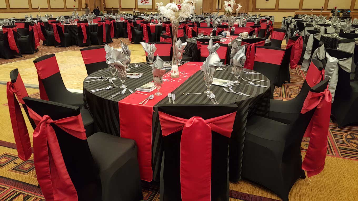Formal banquet hall with black chair covers, red sashes and silver napkin folds, luxury linen décor package by Pizazz Rents & Events
